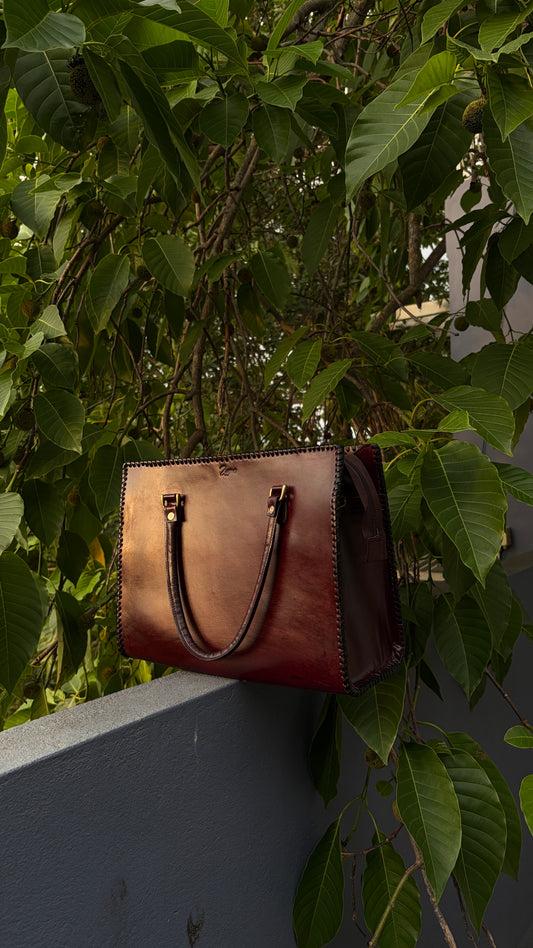 A brown leather handbag with dark brown handles and gold-tone hardware, resting on a gray wall beneath dense green foliage.