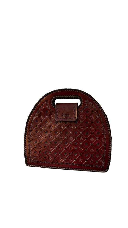 A reddish-brown leather handbag with a quilted diamond pattern and a top handle, resting on a gray ledge surrounded by green leaves.