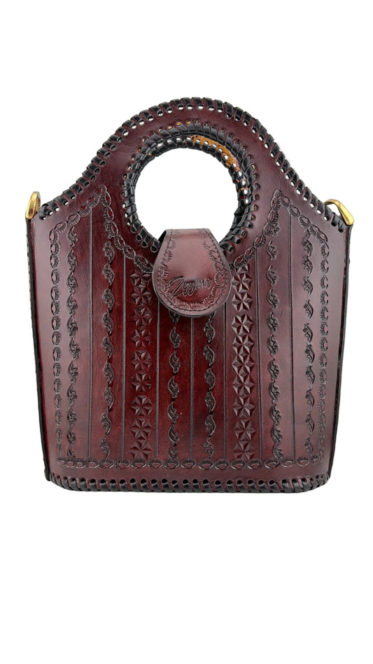 A dark reddish-brown leather handbag with a circular top handle, featuring intricate hand-tooled floral and geometric patterns, a front flap pocket with a leather tab, and gold-tone hardware on a light gray background.