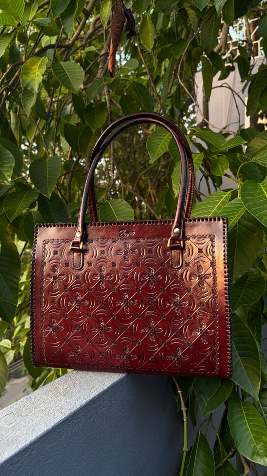 A dark reddish-brown embossed leather handbag with double top handles, resting on a gray ledge surrounded by green leaves.