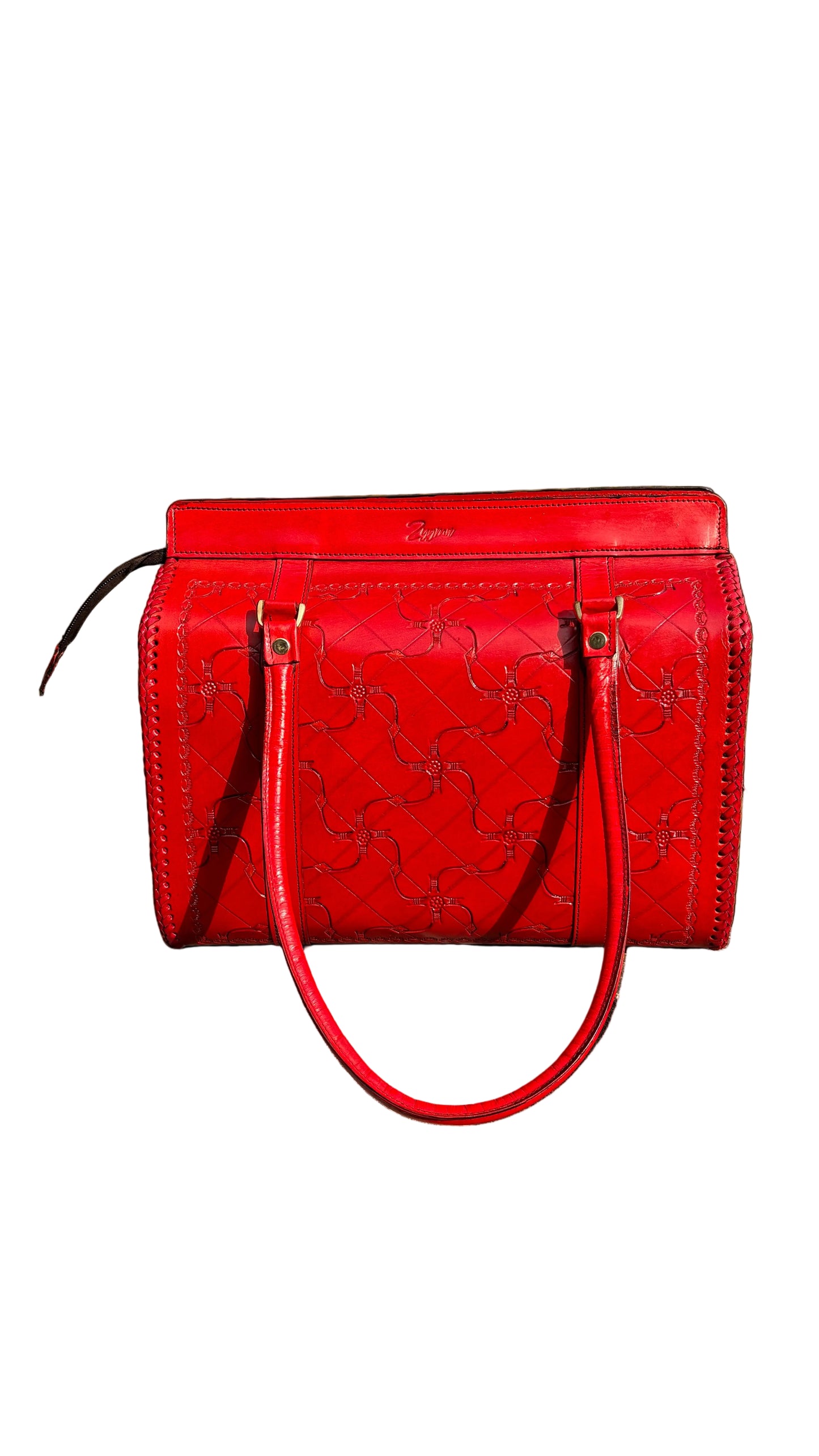 A bright red embossed leather handbag with double top handles, resting on a concrete ledge outdoors with green foliage and a blue sky in the background.