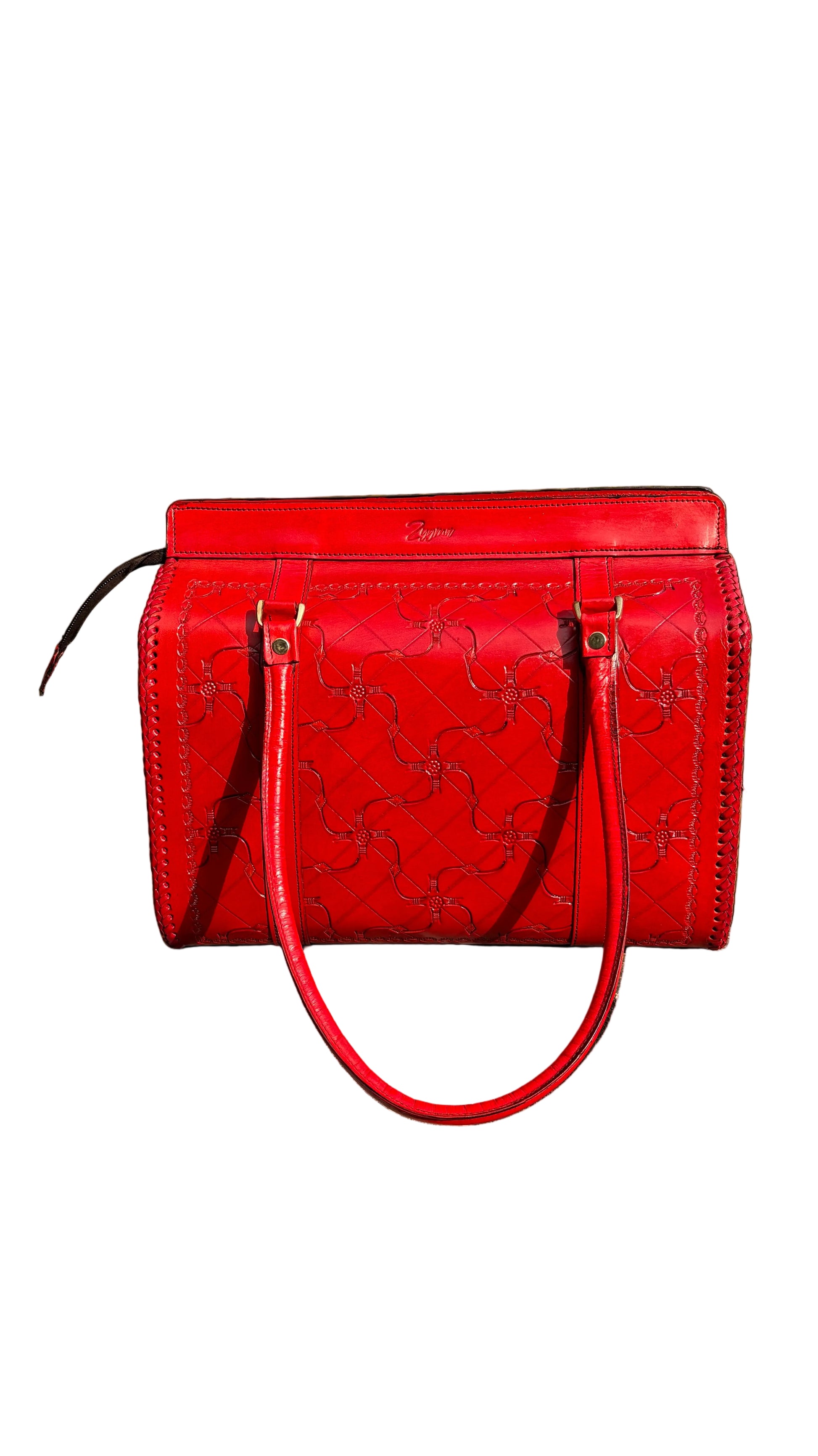 A bright red embossed leather handbag with double top handles, resting on a concrete ledge outdoors with green foliage and a blue sky in the background.