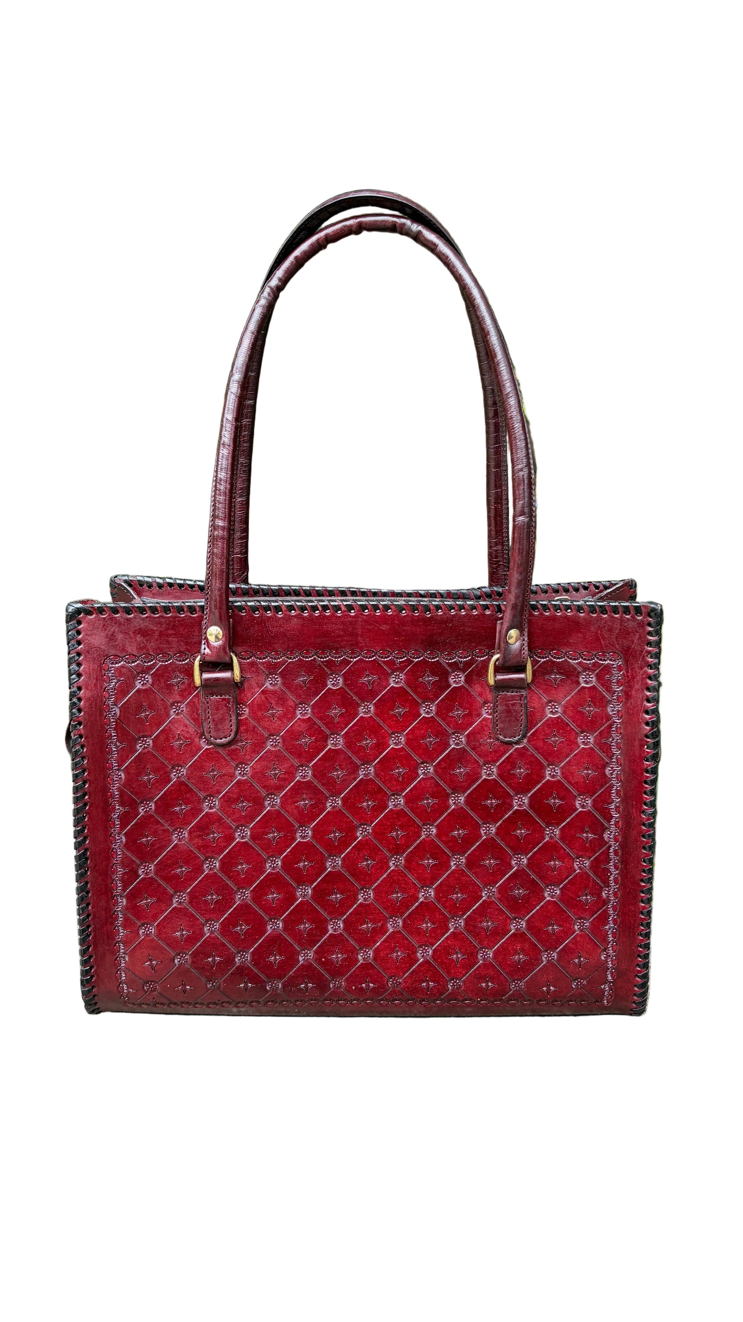 A red leather tote bag with a repeating monogram pattern and double top handles, resting on a concrete ledge with a blurred background of green trees.