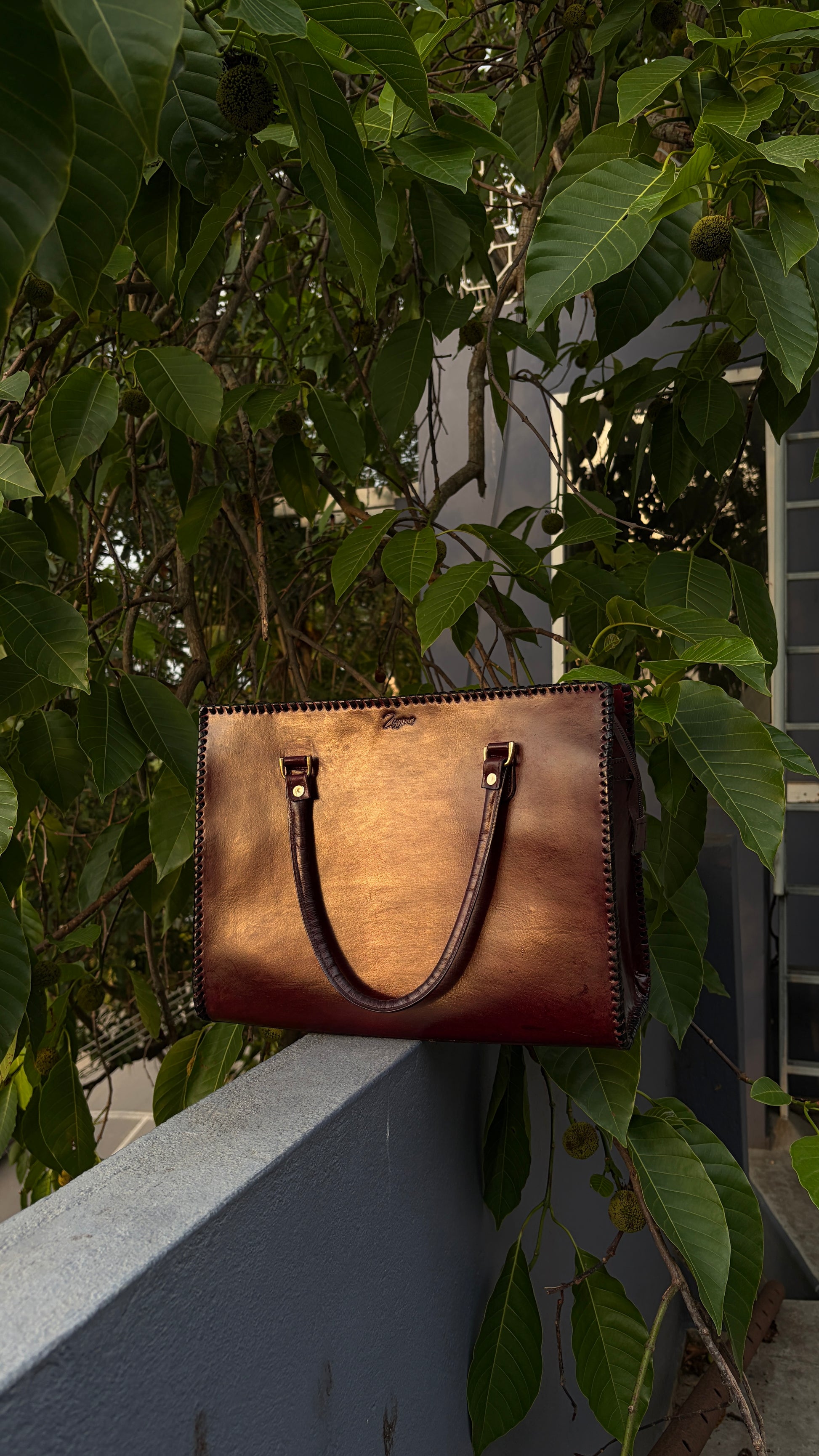 A brown leather tote bag with dark brown handles and gold-tone hardware, resting on a gray concrete ledge surrounded by lush green foliage.