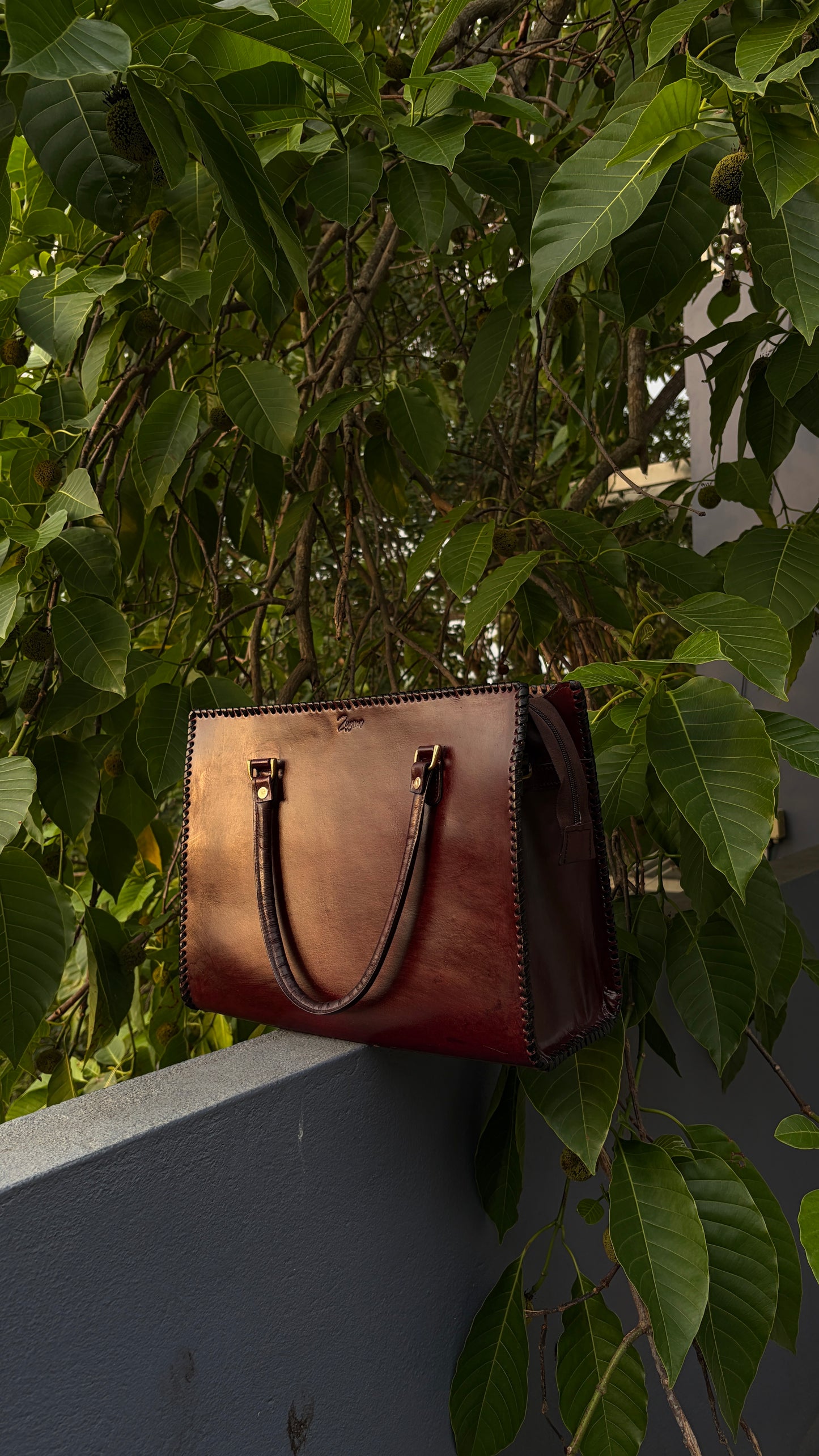 A brown leather handbag with dark brown handles and gold-tone hardware, resting on a gray wall beneath dense green foliage.
