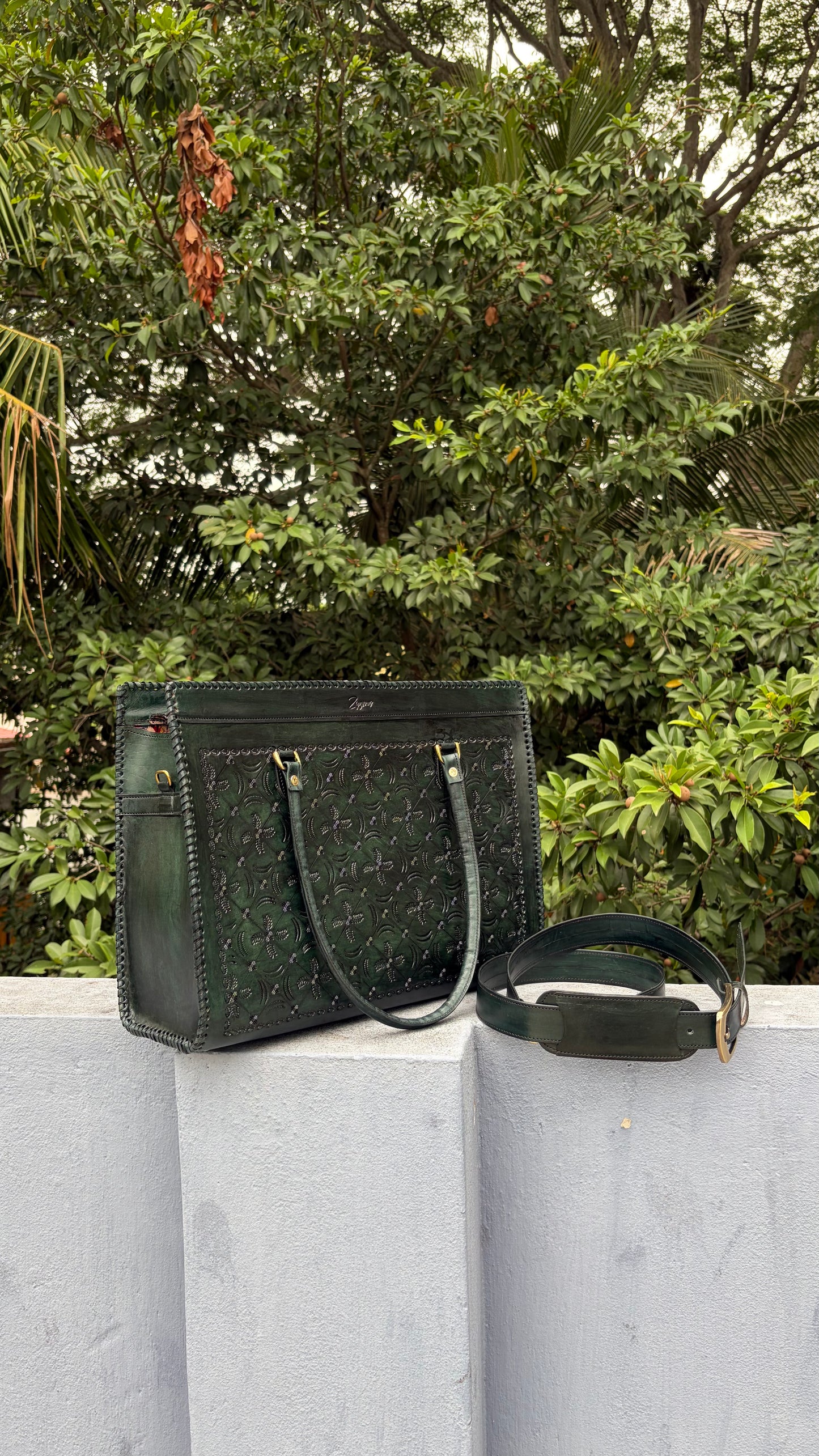 A dark green leather tote bag with an embossed monogram pattern sits on a light gray concrete ledge outdoors, with a matching shoulder strap placed beside it and dense green foliage in the background.